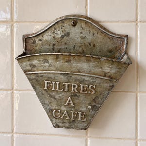 Può includere: Un portafiltri per caffè in metallo da parete con una finitura grigia usurata e invecchiata. La parte anteriore presenta scritte argentate in rilievo che recitano FILTRES A CAFE. Il contenitore ha un'ampia apertura superiore e una piastra.