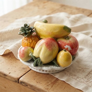 Op de afbeelding: Een keramische fruitschaal gevuld met een decoratief assortiment van realistisch ogend fruit, waaronder een gele banaan, een gestructureerde ananas, een rood-gele perzik, een rode appel en een kleine gele citroen, gerangschikt op een wit bord.