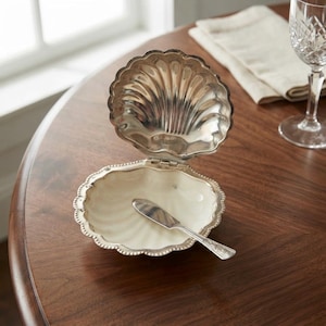 May include: A silver-toned, clam-shaped serving dish with a hinged lid that opens upward. The interior of the dish is smooth and off-white, while the exterior and edges feature a metallic finish. A small, matching silver-toned spreader knife rests inside.