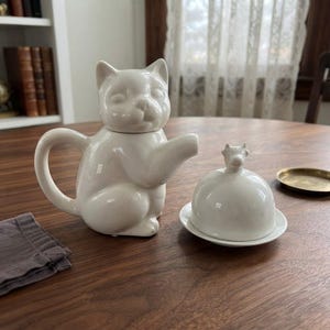 Può includere: Una teiera in ceramica bianca a forma di gatto seduto con manico curvo e beccuccio, accompagnata da un burriera in ceramica bianca abbinata con coperchio a cupola sormontato da una piccola statuetta di animale, finitura liscia e lucida.