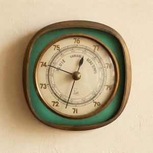 May include: A square-shaped wall barometer with rounded corners, featuring a brass-toned frame and a teal-colored border. The central cream-colored dial displays black numerical markings, the text "PLUIE," "VARIABLE," and "BEAU TEMPS," and two needles.