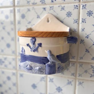 Puede incluir: Un salero de pared de cerámica con temática náutica que presenta un ancla azul, un velero, una gaviota y conchas marinas en relieve. El recipiente tiene una base de color crema claro, una tapa de madera natural y un asa pequeña.