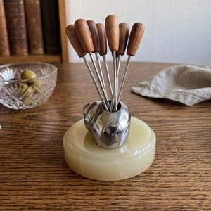 May include: A set of seven stainless steel and wood-handled appetizer forks displayed in a silver-colored holder. The holder sits on a round, light yellow onyx base. Olives in a crystal bowl are visible in the background.