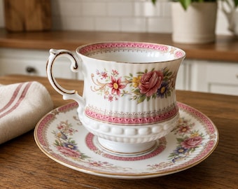 May include: A white porcelain teacup and saucer set featuring a delicate floral pattern with pink, red, and yellow roses. The rim is decorated with a pink border and gold trim. The cup has a fluted base and a curved handle with gold accents.