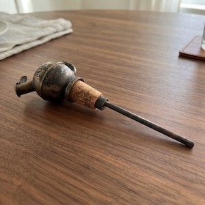 Peut inclure: Un verseur de bouteille en métal vintage doté d'une partie supérieure sphérique à la patine sombre et usée, d'un bouchon en liège naturel et d'un long bec verseur métallique fin. Le design comprend un petit capuchon articulé sur le côté.