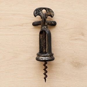 Op de afbeelding: Een vintage metalen kurkentrekker met een donkere, verweerde afwerking met zichtbare tekenen van oxidatie en slijtage. Het gereedschap bevat een centrale spiraalvormige worm, een stevig frame en een handvat aan de bovenzijde met een opening.