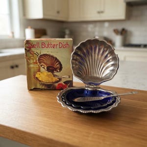 Può includere: Un burriera in metallo argentato a forma di conchiglia con un inserto in vetro blu intenso e un coltello spalmaburro abbinato. L'oggetto è esposto accanto alla sua scatola di cartone originale con la scritta Shell Butter Dish in rosso.