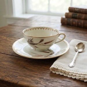 Peut inclure: Une tasse à thé et sa soucoupe en porcelaine crème ornées d'une illustration botanique délicate de tiges de blé brunes. Le bord de la tasse et de la soucoupe est décoré d'un fin liseré doré et d'un motif festonné répétitif en or.