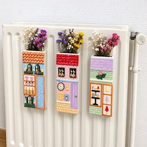 Può includere: Tre vasi da parete rettangolari in ceramica a forma di facciate di negozi colorate, riempiti con fiori secchi. Il vaso a sinistra ha tegole arancioni, quello centrale un orologio e facciata rosa, quello a destra un motivo a mattoni viola.