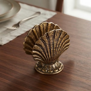 Op de afbeelding: Een metalen, goudkleurige servettenhouder in de vorm van een grote open schelp. Het object heeft een getextureerde, verweerde afwerking met diepe ribbels die vanuit de basis naar de geschulpte randen lopen, staand op een ronde voet.