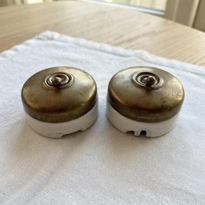 Può includere: Due interruttori della luce rotondi con una copertura superiore in metallo color ottone e una base in ceramica bianca. Ogni interruttore presenta una levetta centrale e la scritta 6A 250V in rilievo sul lato dell'involucro in ottone.