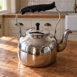 Op de afbeelding: Een theeketel van gepolijst zilverkleurig metaal met een ronde behuizing en verticale ribbels. De ketel is voorzien van een zwart kunststof handvat met vingergrepen, een bijpassende zwarte knop op het afneembare deksel en een gebogen tuit.