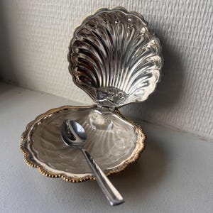 May include: A silver-plated clam shell dish with a hinged lid and a beaded gold-tone rim. The interior features a polished, reflective surface with a scalloped design. A small, matching silver-tone metal spoon rests inside the open shell container.