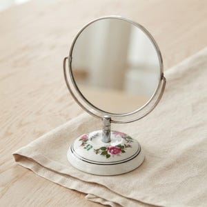 May include: A round, silver-toned metal tabletop mirror mounted on a swivel frame. The base is made of white ceramic and features a decorative floral pattern with pink roses and green leaves, accented by a thin black circular line around the edge.