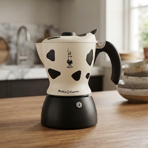 Pode incluir: Uma cafeteira Bialetti Mukka Express com a parte superior em creme com manchas pretas de vaca, uma pega preta e uma base preta sólida. O logótipo Bialetti e o texto Mukka Express estão impressos na parte da frente.