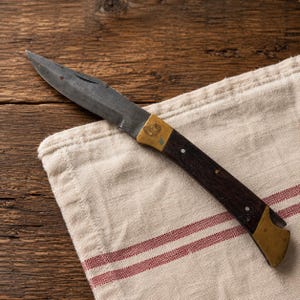 Könnte beinhalten: Ein Klappmesser mit einer dunklen, verwitterten Stahlklinge und einem Griff aus dunklen Holzschalen. Der Griff verfügt über Messingbacken an beiden Enden, ein kleines eingraviertes Emblem auf der oberen Backe und zwei silberne Stifte.