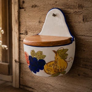 Może przedstawiać: Biały ceramiczny pojemnik ścienny z drewnianą półką i niebieską obwódką. Pojemnik jest ozdobiony malowanym wzorem owoców, w tym winogron i gruszek. Mały metalowy haczyk znajduje się na górze.