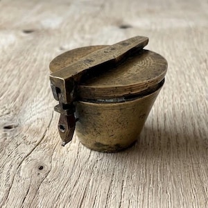 May include: An antique brass container with a hinged lid and a handle. The container has a cylindrical shape with a slightly flared base. The brass has a tarnished, aged appearance, suggesting its vintage. The handle is rectangular and lies flat against the lid.