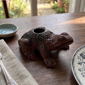 May include: A brown frog-shaped candle holder with a textured surface. The frog has a large opening on its back for a candle. A small blue bowl and a decorative plate are also visible.