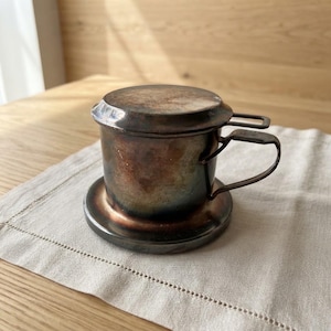 Könnte beinhalten: Ein zylindrischer vietnamesischer Kaffeefilter aus Metall mit einer dunklen, oxidierten Patina, bestehend aus einem flachen Deckel, einem seitlichen Griff und einer runden Bodenplatte. Die Metalloberfläche zeigt gesprenkelte Braun-, Kupfer- und Anthrazittöne.