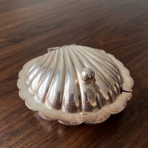 Könnte beinhalten: Eine silberfarbene Schmuckschatulle in Muschelform mit einem kleinen, runden Knopf oben. Die Schatulle hat einen gewellten Rand und einen Klappdeckel. Die Oberfläche reflektiert das Licht, was auf eine polierte Oberfläche hindeutet. Die Schatulle ist wahrscheinlich aus Metall.