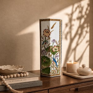 Puede incluir: Un jarrón blanco rectangular con ilustraciones florales y de aves. El jarrón tiene un borde dorado y está sobre una superficie de madera. También se ven una vela encendida y un cuenco decorativo.