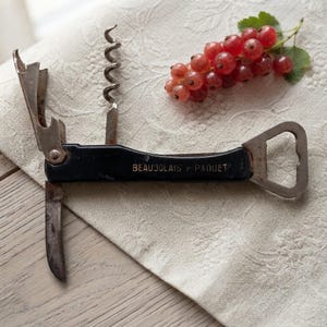 Op de afbeelding: Een zwarte multifunctionele kurkentrekker met een metalen spiraal, een klein mesje en een flesopener, met de tekst BEAUJOLAIS PAQUET op het handvat, vergezeld van een klein trosje rode bessen met groene blaadjes.