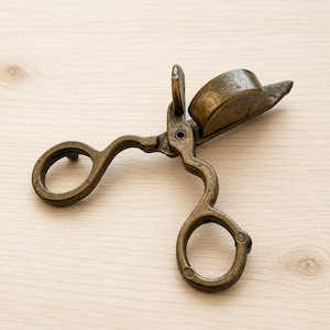 May include: An antique brass candle wick trimmer featuring two circular finger loops and a hinged box mechanism designed to catch trimmed wicks. The metal has a weathered, dark patina with visible signs of age and a textured, metallic finish.