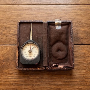 Peut inclure: Un tensiomètre vintage avec un boîtier noir et un cadran circulaire blanc marqué de chiffres de 25 à 150 et du mot GRAMMES. L'instrument possède une fine aiguille métallique et est logé dans un étui rectangulaire marron avec une doublure en velours.