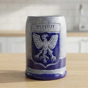 May include: A cylindrical ceramic beer stein featuring a textured grey upper section and a smooth deep blue base. A central shield emblem displays a raised white eagle graphic with the word OBERNAI embossed in capital letters above the bird.