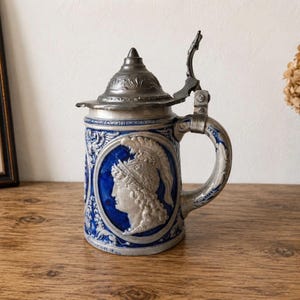 Op de afbeelding: Een blauwe en grijze keramische bierpul met een reliëfportret van een figuur met een gepluimde helm in een centraal ovaal medaillon. De pul heeft een scharnierend metalen deksel met een decoratieve knop en een gebogen handvat.
