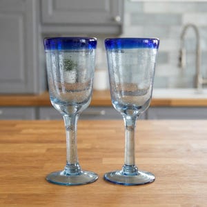 Puede incluir: Dos copas de cristal transparente con un borde azul cobalto distintivo. Cada recipiente presenta un tallo alto y delgado y una base circular ancha. El material de vidrio tiene una apariencia texturizada sutil con pequeñas burbujas de aire.