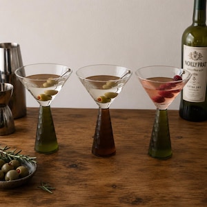 Peut inclure: Trois verres à martini avec des pieds coniques texturés en verre vert et ambre. Deux verres contiennent un liquide clair avec des olives vertes sur des piques en métal, tandis que le troisième contient un liquide rose avec trois framboises.