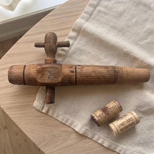 Puede incluir: Un grifo de barril de madera vintage con un mango en forma de T y una boquilla cónica, acompañado de dos corchos cilíndricos. Un corcho tiene impresa la palabra CHATEAU y el otro tiene las palabras VINEYARD RESERVE en tinta negra.
