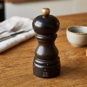 Puede incluir: Un molinillo de pimienta Peugeot de madera marrón oscuro con una parte superior dorada. El molinillo tiene un cuerpo redondo y un acabado texturizado. El logotipo de Peugeot es visible en la base. Un pequeño cuenco de cerámica y una servilleta blanca están en el fondo.
