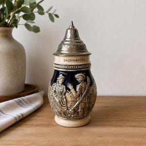 Può includere: Un boccale da birra in ceramica con coperchio conico in metallo, caratterizzato da un corpo blu scuro con rilievi color beige raffiguranti due figure e un cane. Il testo BADEN-BADEN è stampato su una fascia beige sotto il coperchio.