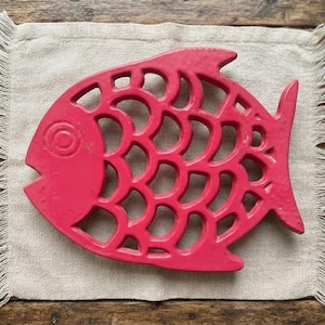 May include: A bright pink cast iron trivet shaped like a fish with a detailed scale pattern and a circular eye. The object features a cut-out design throughout the body, creating a series of interconnected openings that form the fish's skeletal structure.