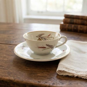 Könnte beinhalten: Eine weiße Teetasse aus Keramik mit passender Untertasse, verziert mit einer zarten botanischen Illustration von Weizenhalmen in Braun-, Grün- und Rottönen. Die Tasse hat einen geschwungenen Henkel und einen glatten Rand auf einer Untertasse.