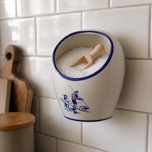 Op de afbeelding: Een aan de muur gemonteerd keramieken zoutvat in een crèmekleurige afwerking met een blauwe rand. De voorzijde toont een blauwe en rode afbeelding van een vissend persoon. De houder is gevuld met wit zout en bevat een klein houten schepje.
