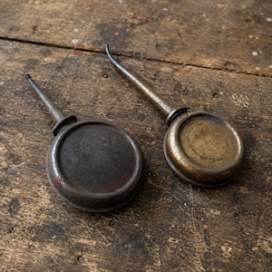 May include: Two vintage metal oil cans with circular bodies and long, tapered spouts. One oil can features a dark, weathered grey finish, while the other displays a tarnished brass-toned patina. Both tools have a simple, functional design.