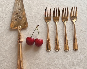 May include: A gold-toned metal cake server with an ornate cutout handle and blade, accompanied by four matching small dessert forks and two fresh red cherries with stems, all arranged together on a textured off-white fabric surface.