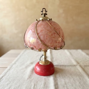 Peut inclure: Une lampe de table dotée d'un abat-jour en verre rose marbré en forme de dôme, entouré d'une armature en fil de laiton. La lampe possède un col en laiton poli et une base rouge circulaire et solide. Un petit fleuron décoratif surmonte le dôme.