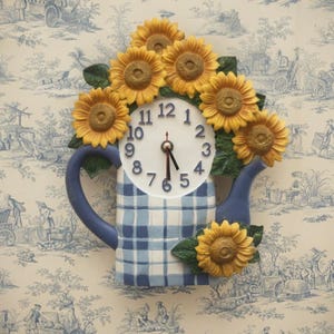 May include: A wall clock shaped like a blue and white chequered watering can, featuring a round white clock face with black numbers and hands. The watering can is decorated with several three-dimensional yellow sunflowers and green leaves.
