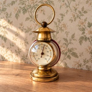 Peut inclure: Un thermomètre en laiton avec un boîtier en verre sphérique et une poignée circulaire sur le dessus. Le cadran blanc présente des graduations numériques noires de -20 à 60 degrés Celsius, avec une aiguille centrale indiquant la température.