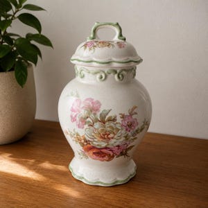 Op de afbeelding: Een crèmekleurige keramische pot met bijpassend deksel met handvat. De pot heeft een craquelé glazuurafwerking, een geschulpte voet en een decoratieve groene rand. Het is versierd met een bloemmotief in roze, oranje en groene tinten.