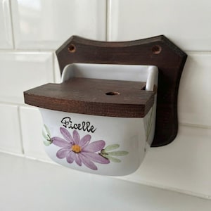 May include: A wall-mounted string holder featuring a white ceramic container with a purple flower illustration and the word Ficelle printed in black. The unit includes a dark brown wooden backplate and a matching wooden lid with a small circular hole.
