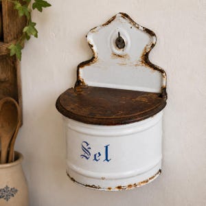 Puede incluir: Una caja de sal de metal esmaltado blanco para montar en la pared con una tapa de madera marrón oscuro. El frente presenta el texto azul "Sel" en letra gótica. La pieza tiene una placa trasera decorativa con un orificio y óxido en los bordes.