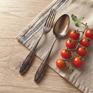 Puede incluir: Un juego de tenedor y cuchara de metal plateado de estilo vintage con mangos oscuros y ornamentados con detalles intrincados, colocado junto a un racimo de tomates cherry rojos frescos en rama sobre una servilleta de tela beige texturizada.