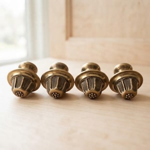 May include: Four brass-toned metal cabinet knobs arranged in a horizontal row. Each knob features a wide, smooth circular base, a fluted, bell-shaped body with vertical ridges, and a small, flat circular top decorated with a cluster of raised dots.