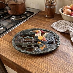 May include: Oval cast iron trivet featuring a colorful rooster design. The rooster has red, yellow, and blue accents. The trivet is designed with a fence and foliage. A decorative kitchen item.